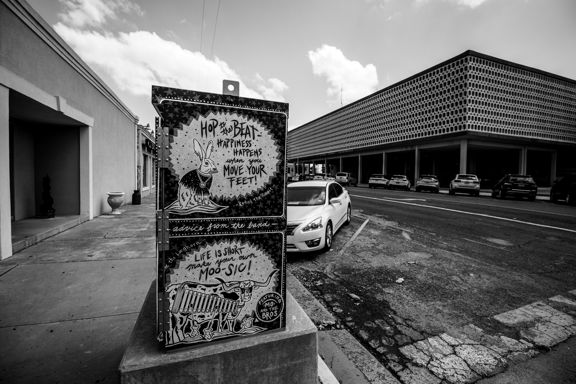 A decorated electrical box with the words "Hop to the Beat - Happiness happens when you move your feet" and "Life is short. Make your own moo-sic" in a downtown area